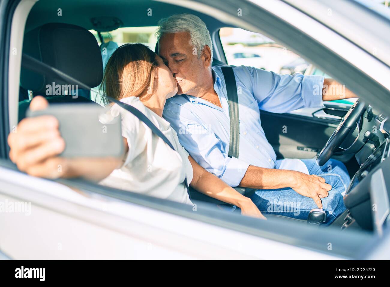 Couple d'âge moyen amoureux assis à l'intérieur de la voiture allant pour un voyage prenant une photo de selfie avec un smartphone embrassant Banque D'Images