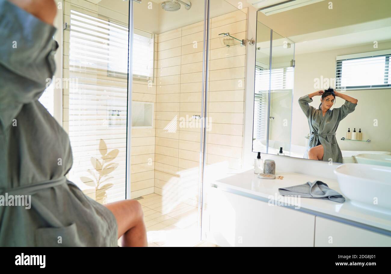 Femme en peignoir fixant les cheveux dans le miroir de salle de bains ensoleillé Banque D'Images