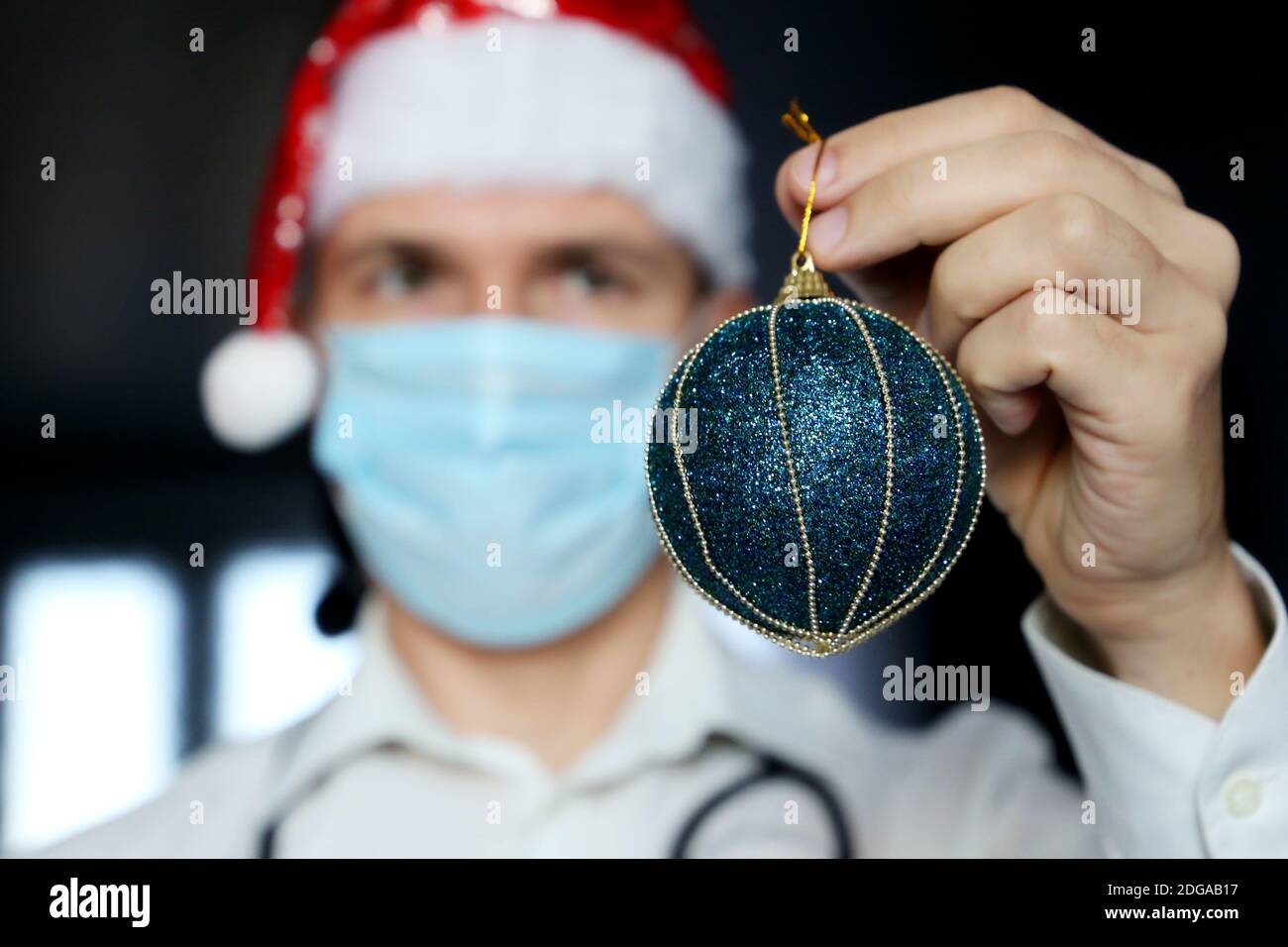 Fête de Noël, docteur en masque et chapeau de père Noël avec une balle de jouet bleue à la main. Concept de décoration de clinique ou de bureau pour les vacances du nouvel an Banque D'Images