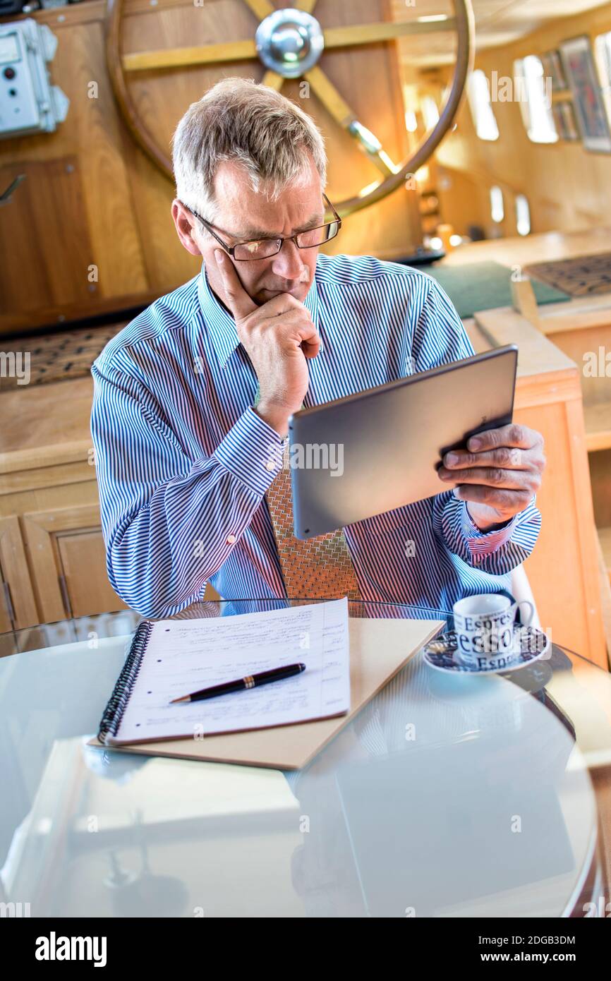 HOMME D'AFFAIRES iPad Zoom/Equipes/FaceTime réunion en cours, homme d'affaires mature se concentrant dans son bureau de barge de bateau tenant une tablette intelligente Apple iPad ordinateur dans une situation de réunion virtuelle Banque D'Images