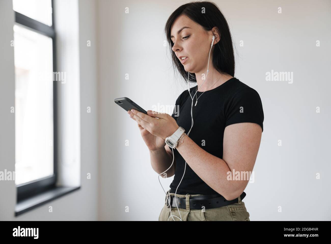 Jeune femme attrayante portant des écouteurs debout à la fenêtre, tenant un téléphone portable Banque D'Images