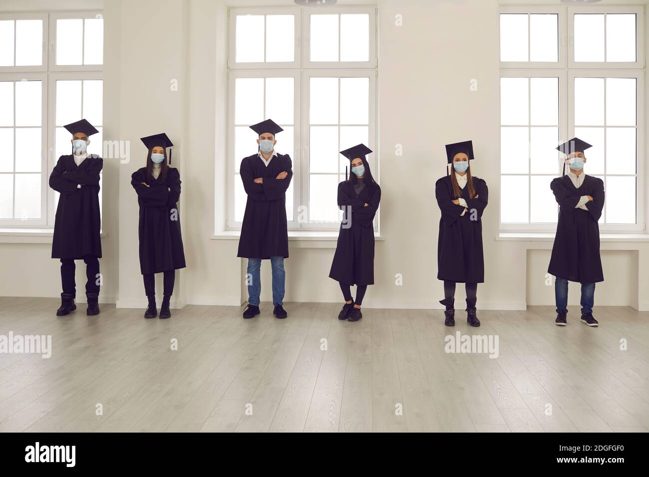 Les étudiants en robes de remise des diplômes et masques de visage debout dans la salle d'université, gardant la distance sociale Banque D'Images