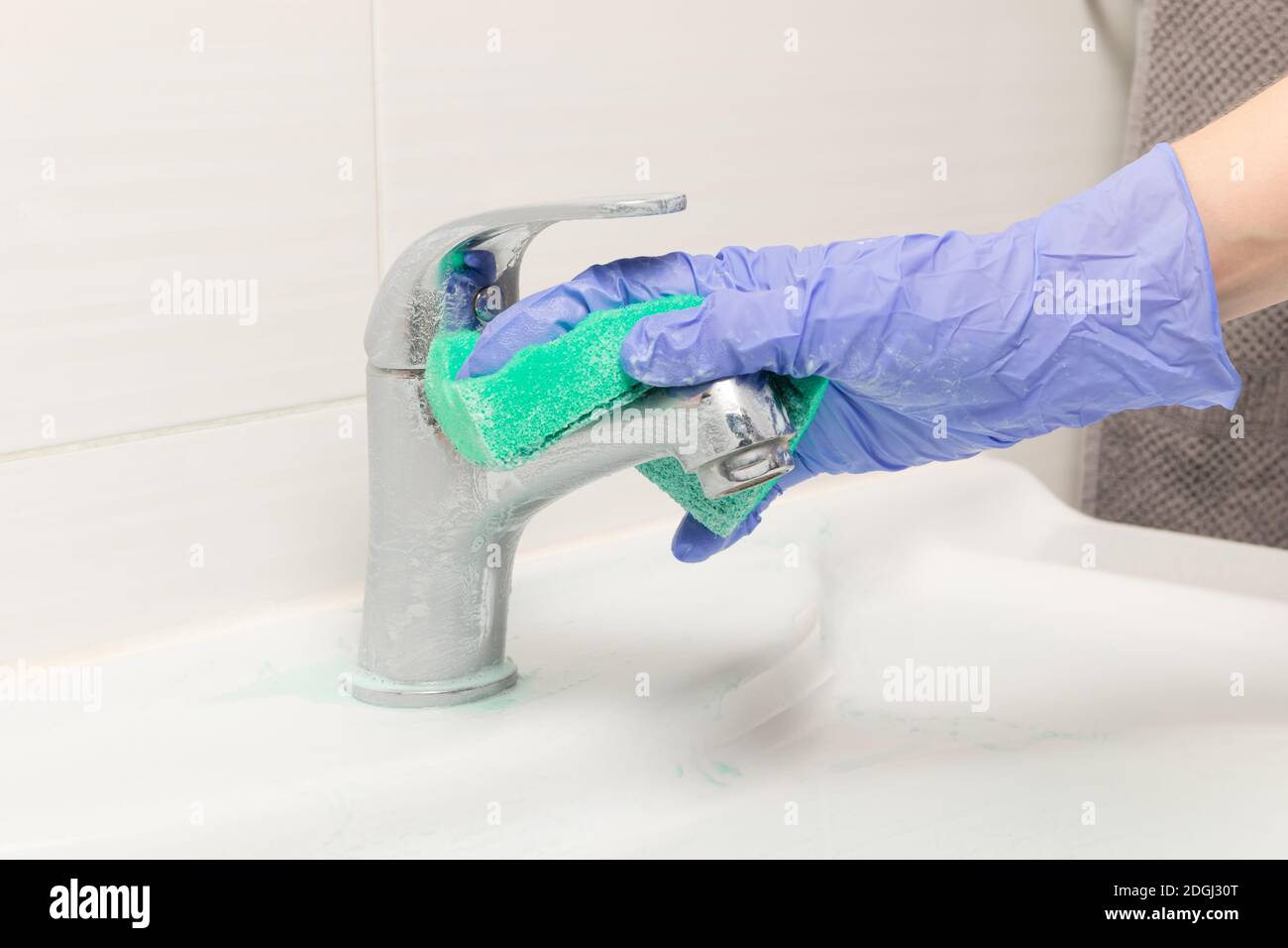 une main femelle dans un gant bleu essuie le mélangeur avec une éponge en mousse verte dans la salle de bains. Nettoyage de la maison Banque D'Images