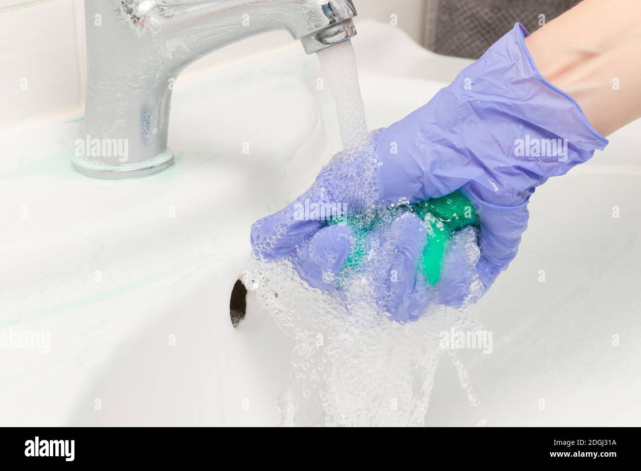 main féminine dans un gant bleu lave une éponge en caoutchouc mousse verte sous un robinet dans la salle de bains. Nettoyage de la maison Banque D'Images