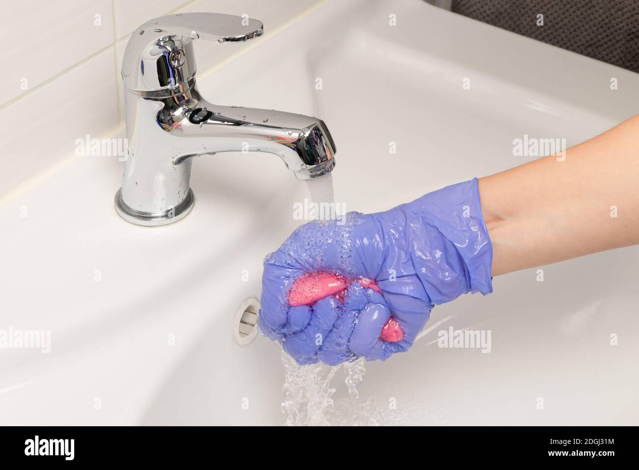 main féminine dans un gant bleu lave un chiffon sous le robinet d'eau dans la salle de bains. Nettoyage de la maison Banque D'Images
