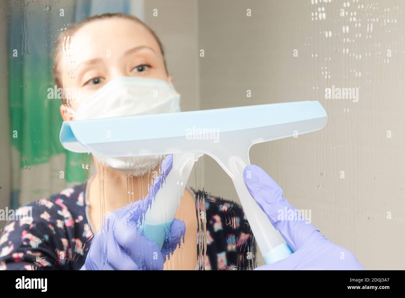 jolie jeune fille femme de ménage dans le masque médical nettoie le miroir de salle de bains au cours d'une pandémie de coronavirus Banque D'Images