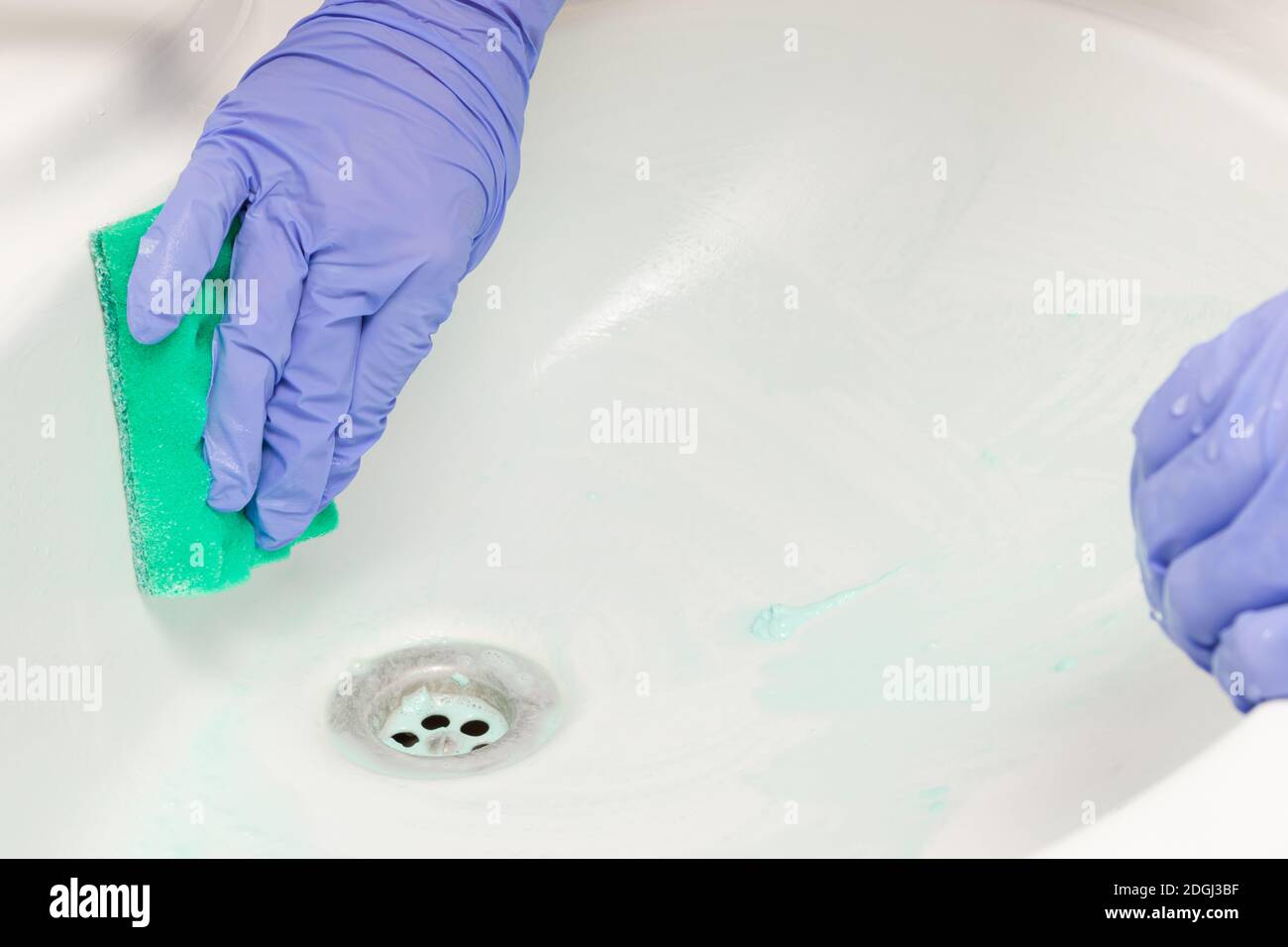une main femelle dans un gant bleu essuie l'évier avec une éponge en mousse verte dans la salle de bains. Nettoyage de la maison Banque D'Images