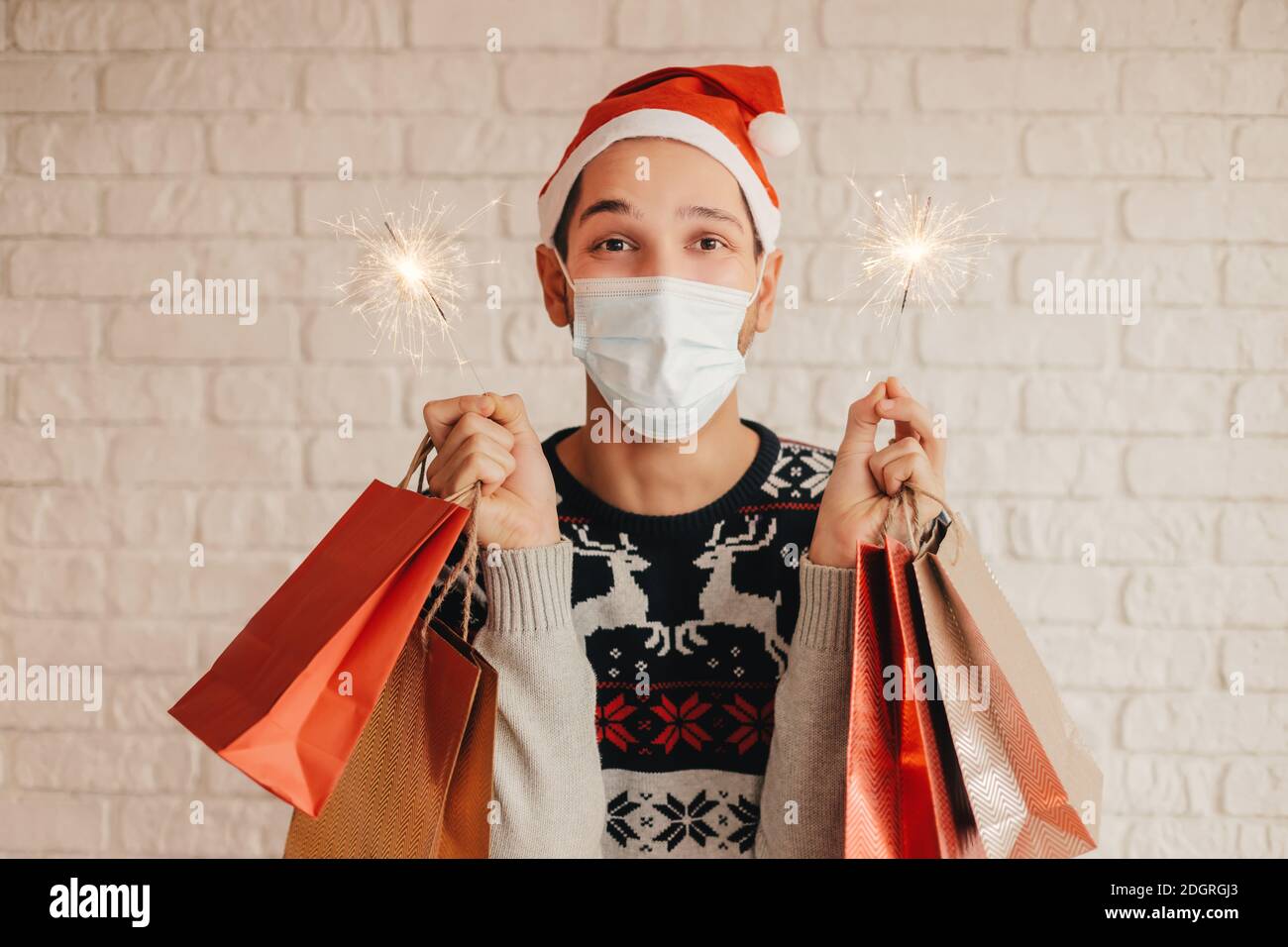 Joyeux jeune homme en chandail de Noël, masque médical pour le visage tenir des sacs d'achats et de la lumière de sparkler dans les mains et le sourire. Joyeux boxeur festif à Sant Banque D'Images