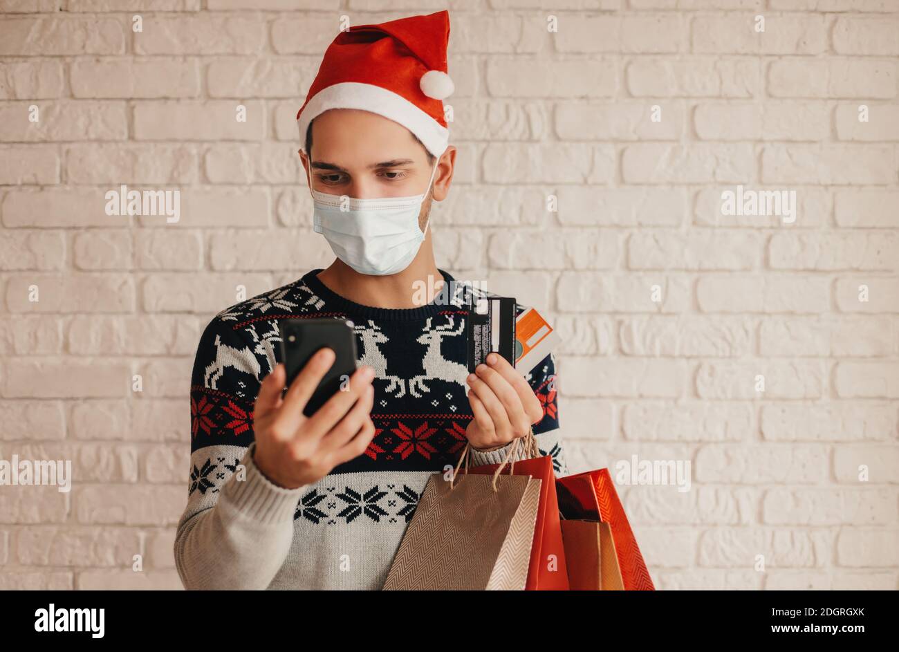 Jeune homme shopper en chapeau de père Noël, masque médical utiliser le smartphone pour le paiement bancaire shopping en ligne. Joyeux homme dans le chandail de Noël, masque de maintien SH Banque D'Images