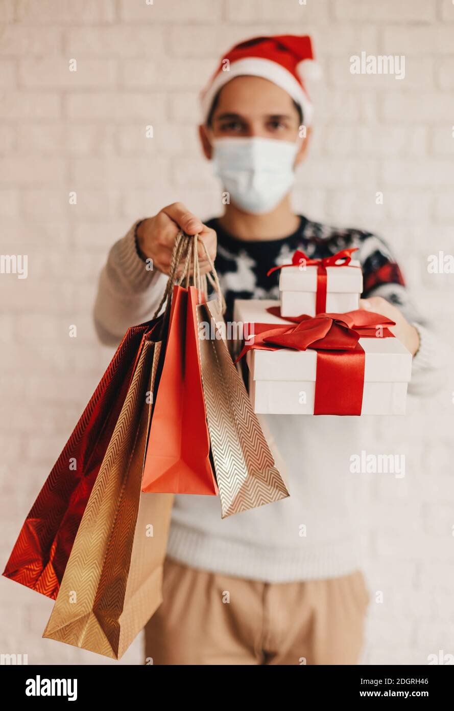 Happy man courier dans le chapeau de Santa, masque médical visage donner des sacs d'achats et des cadeaux de Noël au client. Jeune liveur dans le masque de visage tenir le papier ba Banque D'Images