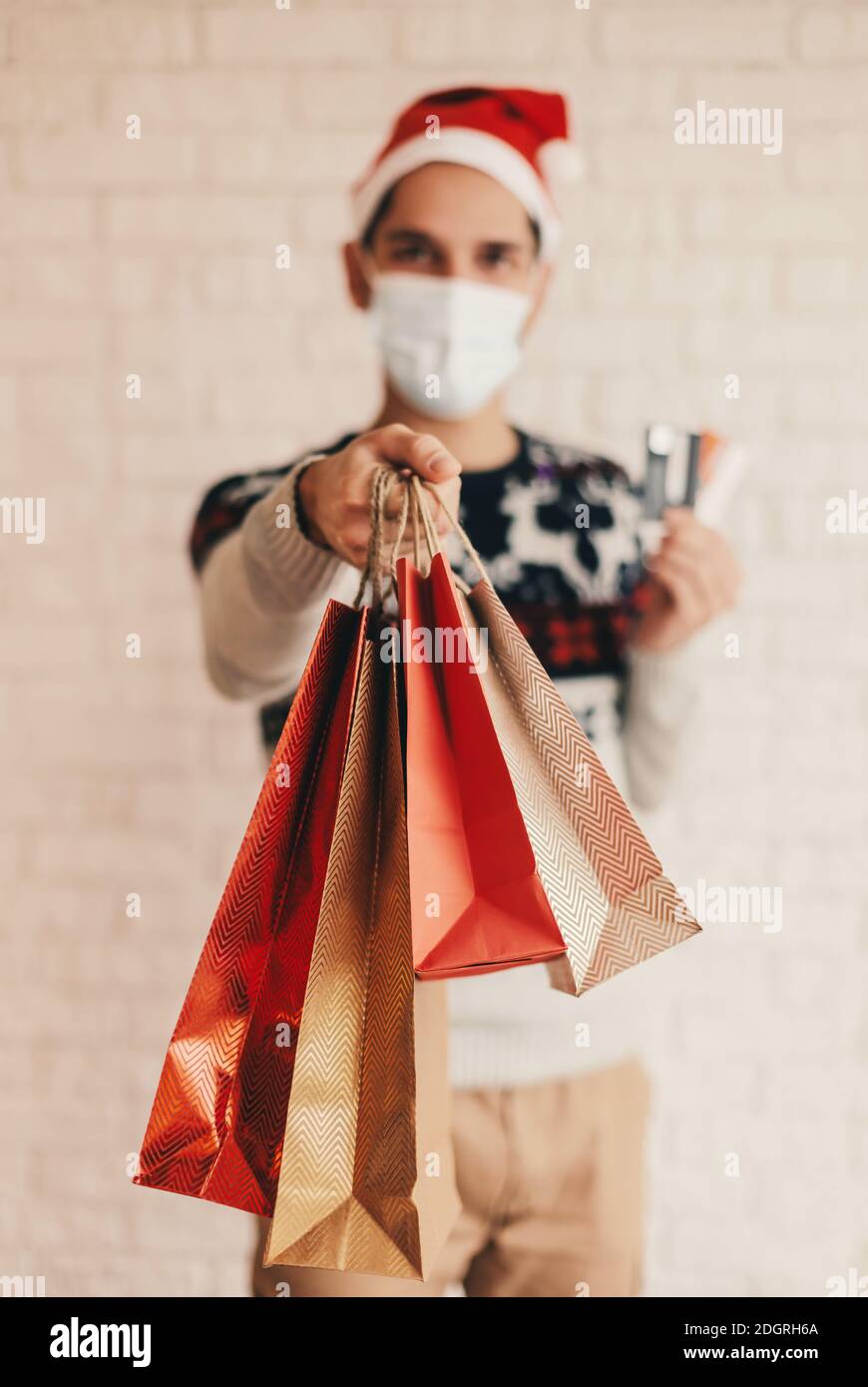 Jeune liveur à chapeau de Santa, masque médical visage donner des sacs d'achats au client. Happy man courier à la caisse du chandail de Noël avec carte de crédit pour Banque D'Images