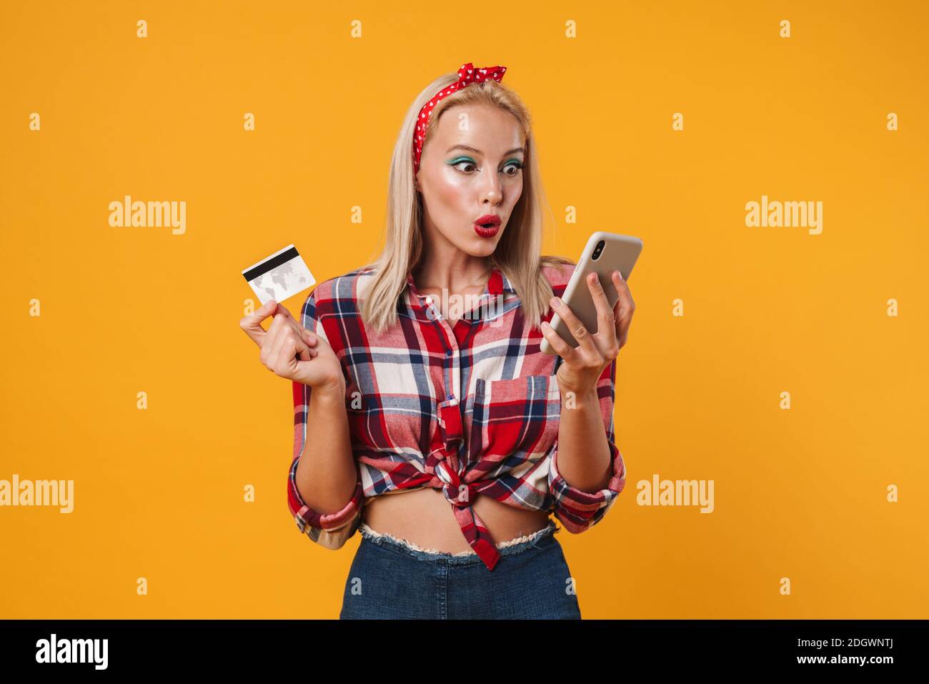 Image de la fille pinup blonde choquée posant avec la carte de crédit et téléphone portable isolé sur fond jaune Banque D'Images