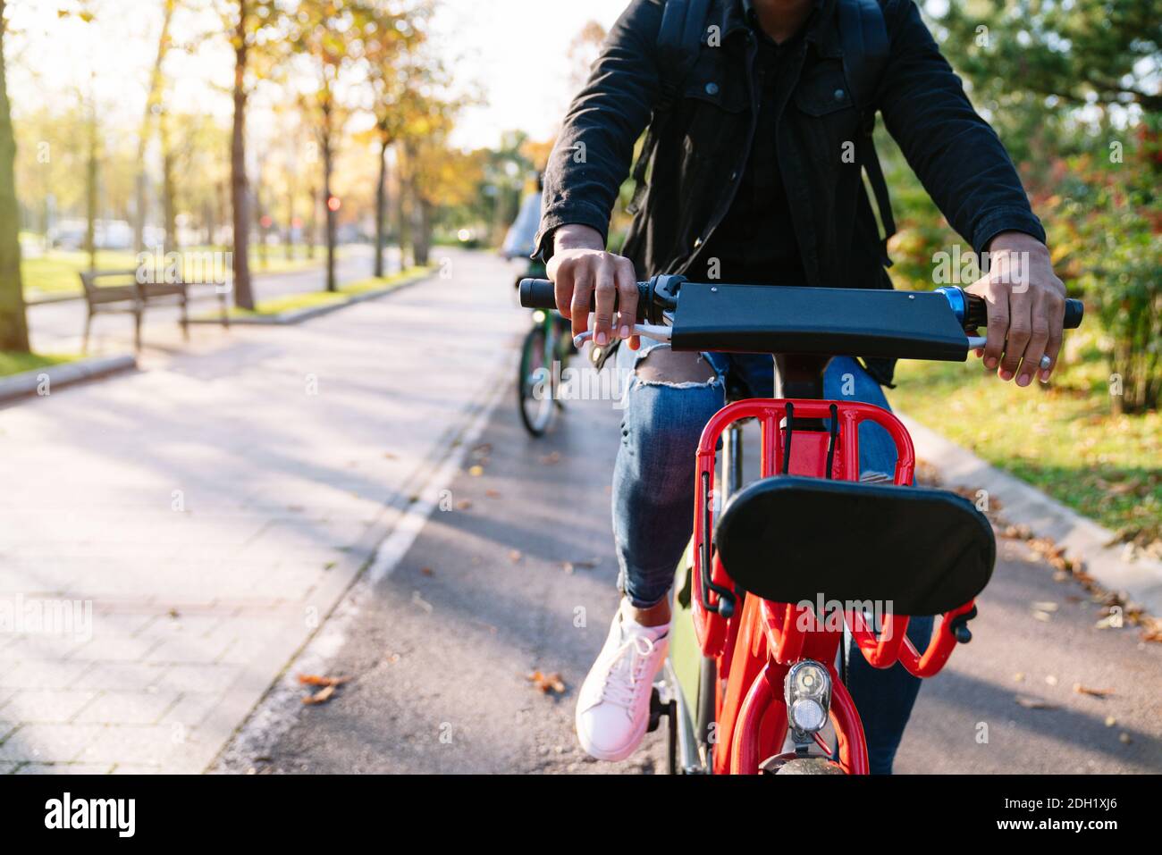 Gros plan photographique du corps d'un garçon étudiant avec un sac à dos sur son dos portant un vélo électrique rouge partagé un sentier de parc bordé d'arbres au coucher du soleil Banque D'Images