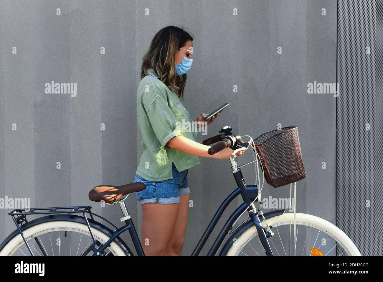 Jeune fille portant un masque facial avec son smartphone tout en tenant un vélo de taille basse sur un fond gris Banque D'Images