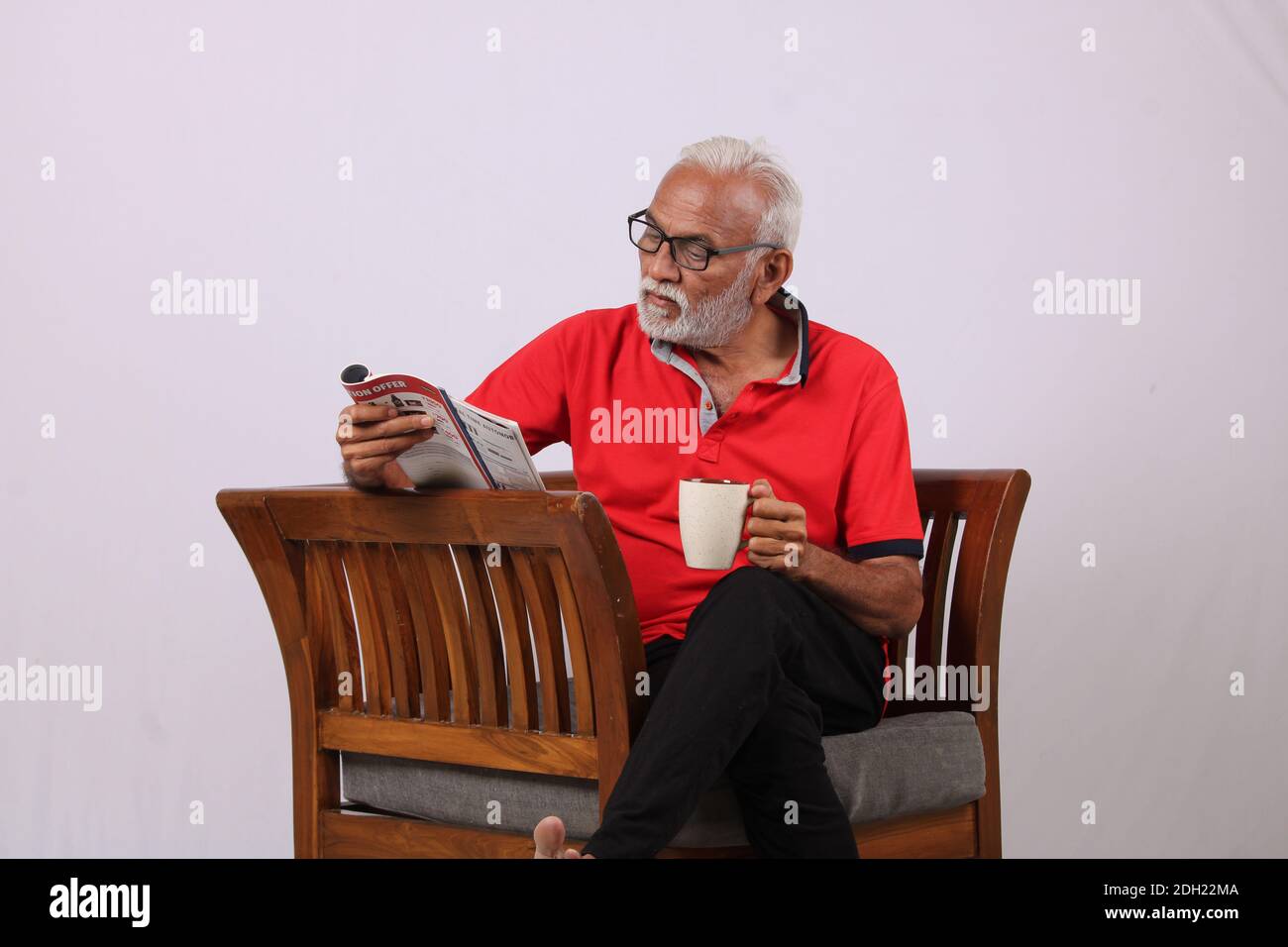 Un vieil homme indien lisant le magazine tout en étant assis sur une chaise avec une tasse de café. Banque D'Images