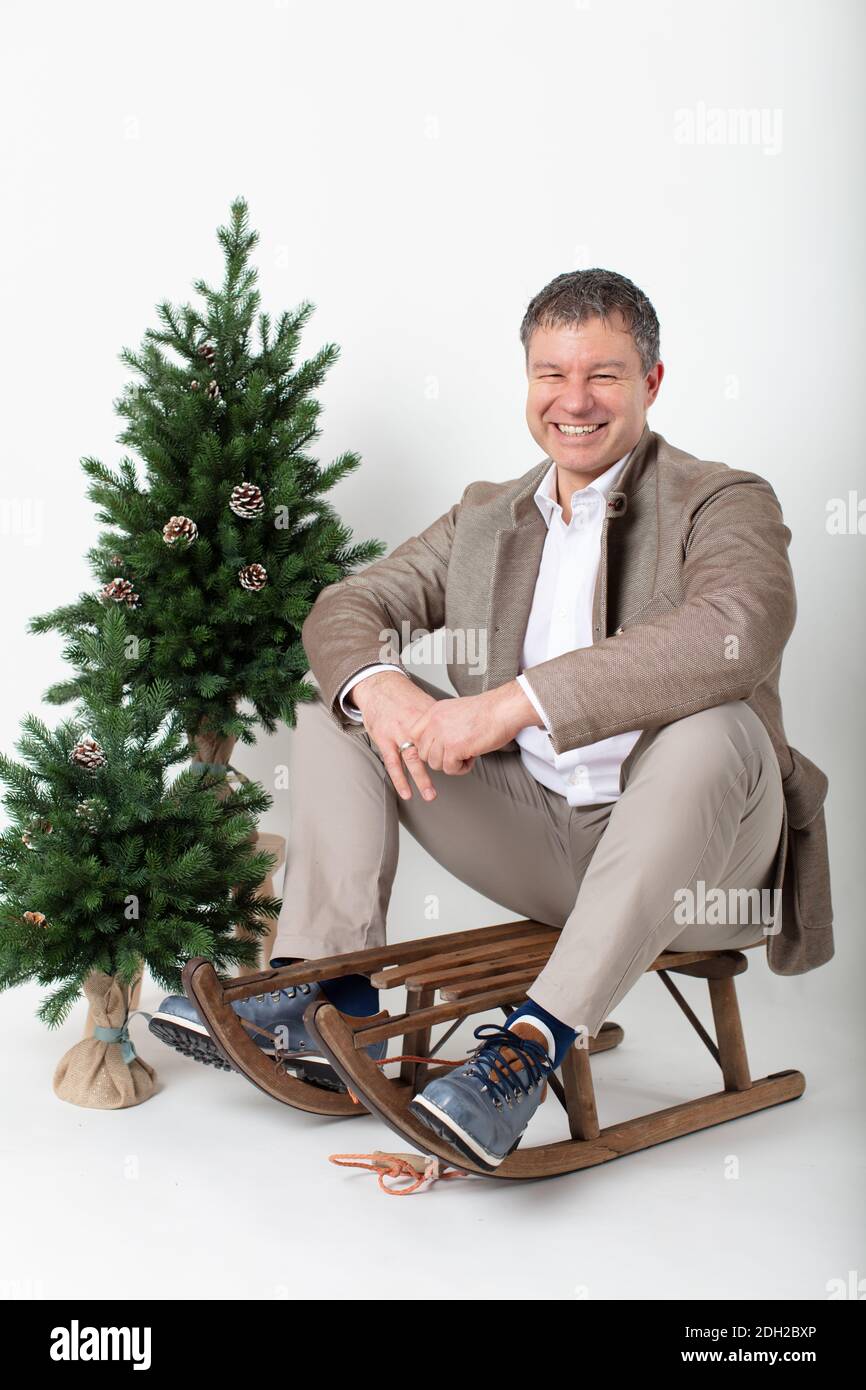 Portrait d'affaires horizontal sur le thème de Noël d'un homme d'affaires habillé élégant et décontracté, assis sur un traîneau en bois rustique sur un fond blanc. Banque D'Images
