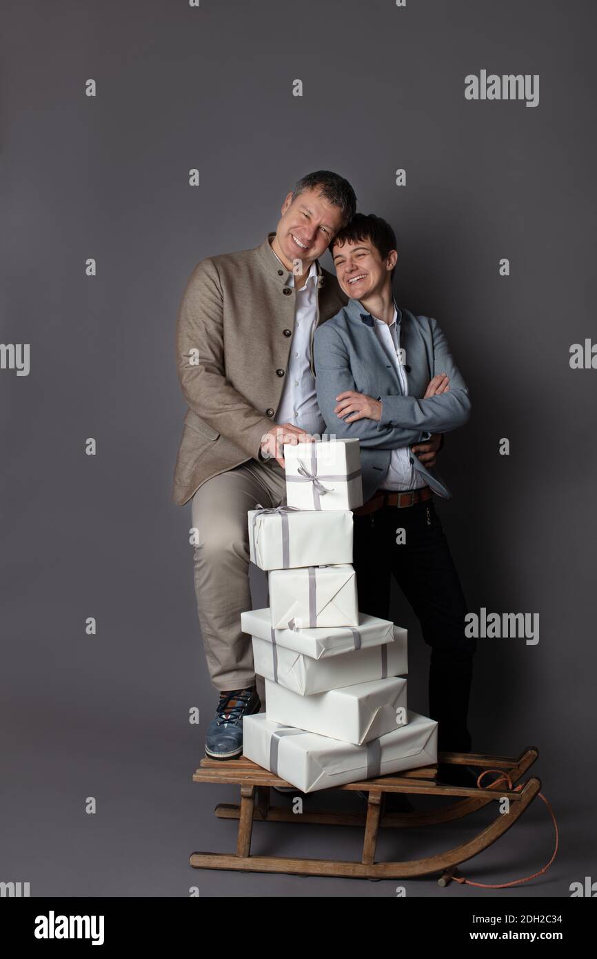 Thème de Noël vertical couple portrait habillé élégant décontracté dans bleu, blanc, beige pantalon et blazers sur fond gris. Au premier plan un traîneau en bois avec une pile de boîtes-cadeaux blanches. Banque D'Images