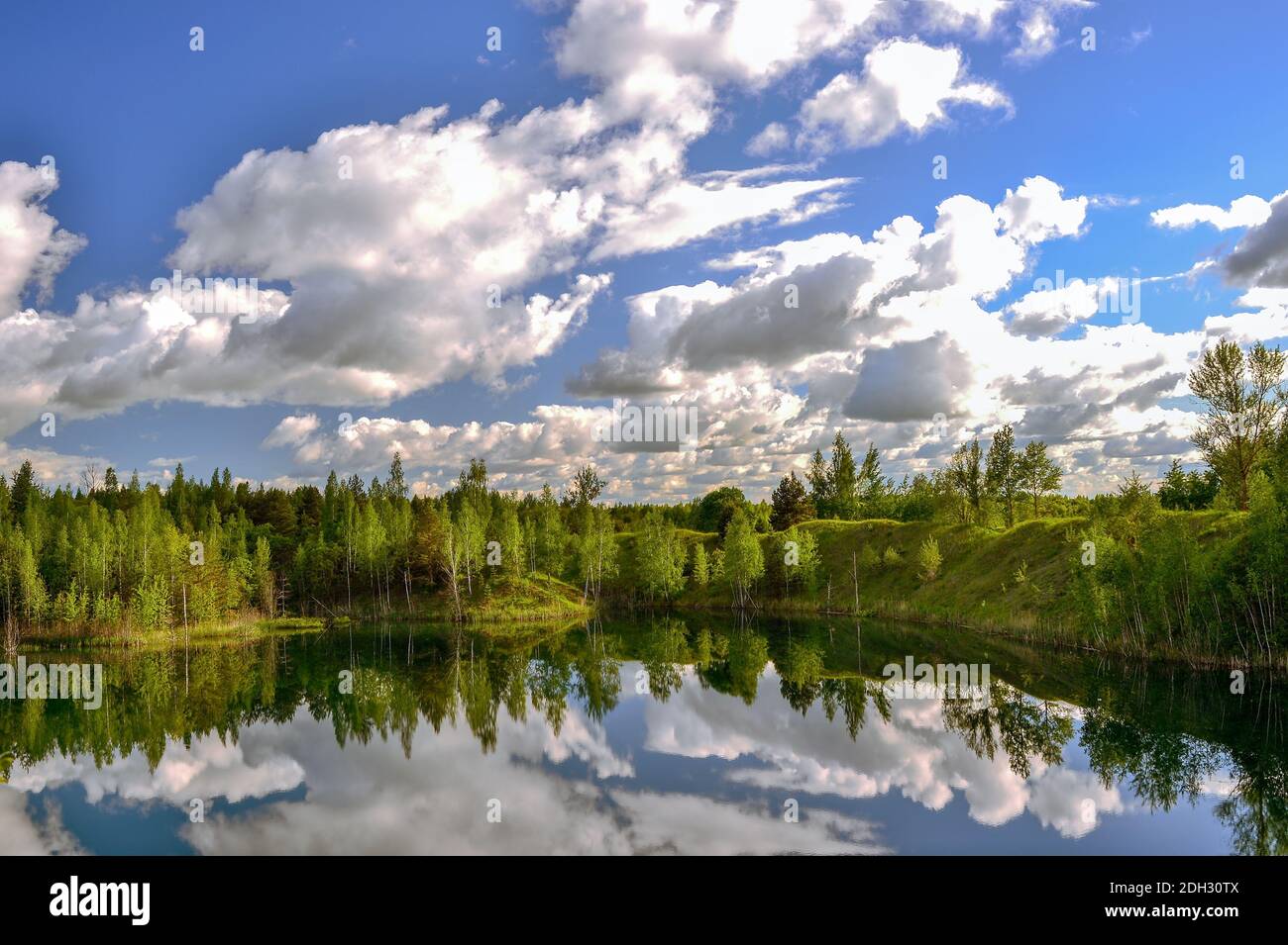 Lac avec des berges dans le vert de printemps brillant de la forêt avec un reflet du ciel bleu avec des nuages dans la surface de l'eau. Arrière-plan. Banque D'Images