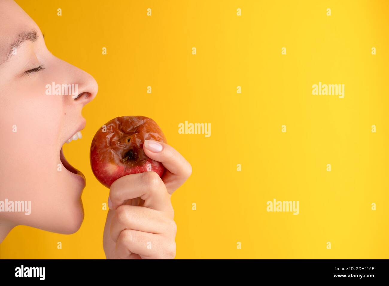 La jeune fille mord une pomme pourrie avec un ver sur fond jaune. Produits périmés, aliments indésirables. Banque D'Images