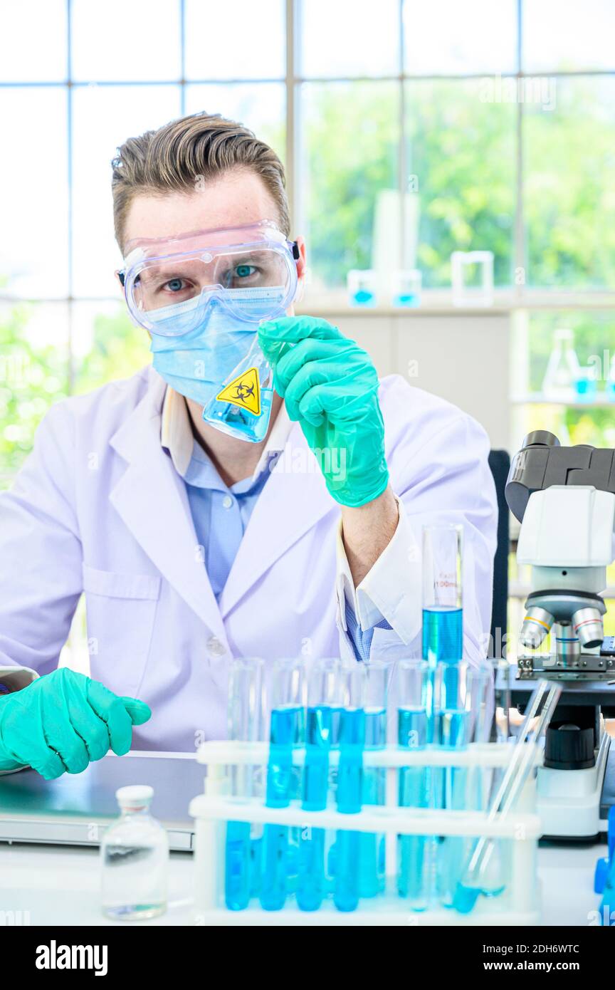 Un scientifique mâle porte une combinaison de protection pour le maintien d'un bécher ou d'un tube avec prélever du liquide à l'intérieur et travailler avec de nombreux équipements de laboratoire pour recherche en laboratoire Banque D'Images