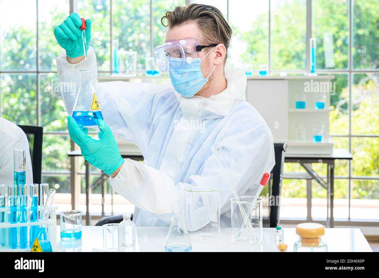 Un scientifique mâle porte une combinaison de protection pour le maintien d'un bécher ou d'un tube avec prélever du liquide à l'intérieur et travailler avec de nombreux équipements de laboratoire pour recherche en laboratoire Banque D'Images