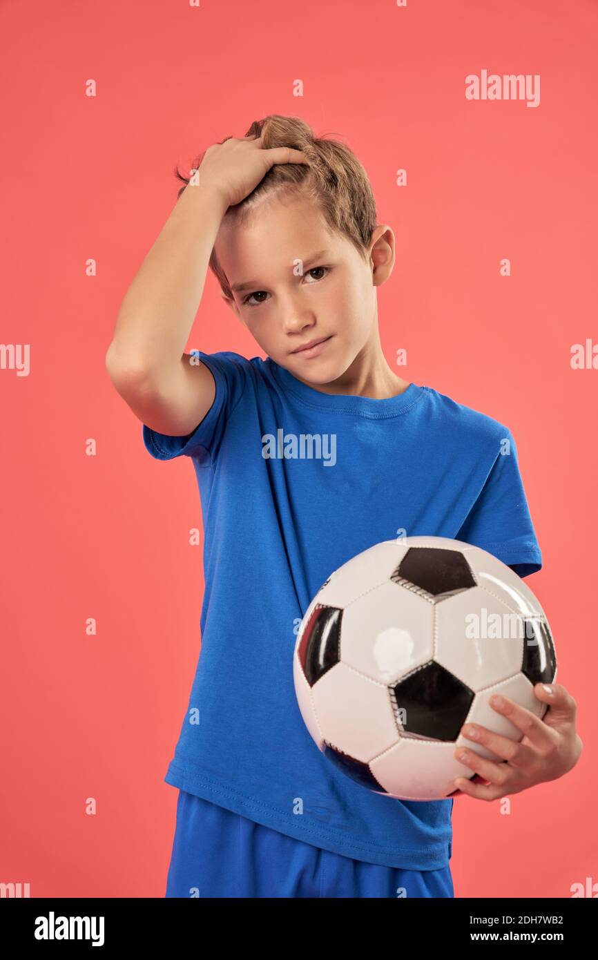 Adorable enfant de sexe masculin en chemise bleue tenant le ballon de football et fixer les cheveux tout en regardant la caméra avec une expression sérieuse Banque D'Images