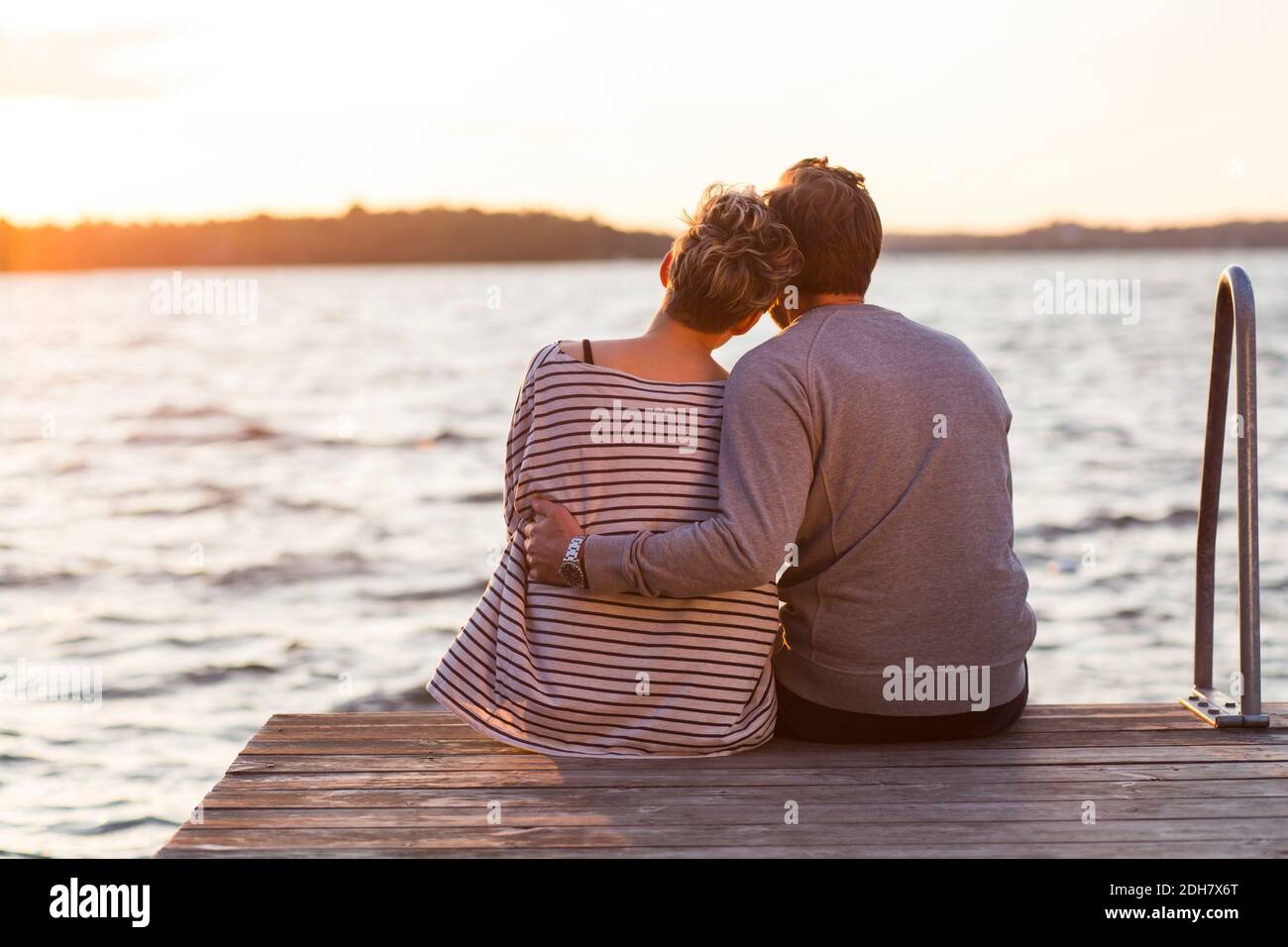 Vue arrière d'un couple affectueux assis sur la jetée contre la mer au coucher du soleil Banque D'Images