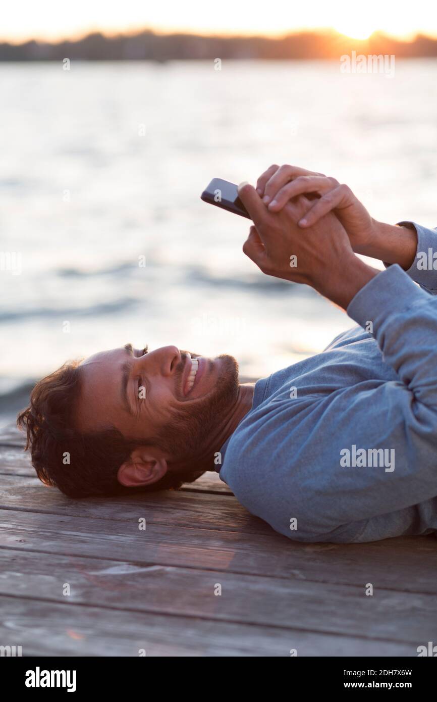 Homme heureux utilisant un téléphone portable en étant allongé sur une jetée en bois contre la mer au coucher du soleil Banque D'Images