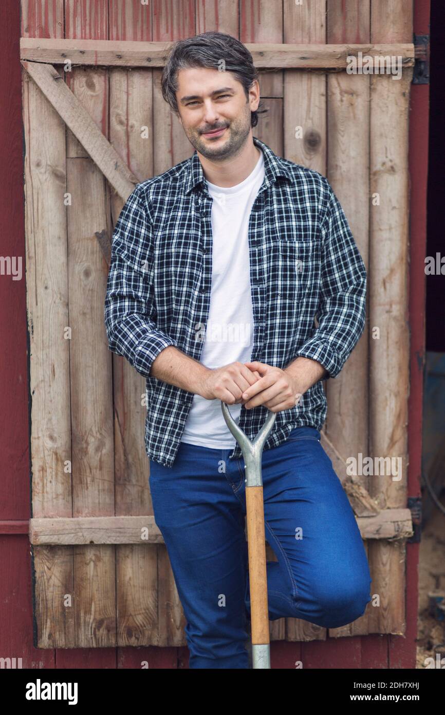 Portrait d'un fermier mâle confiant tenant une bêche tout en se tenant contre porte en bois fermée Banque D'Images