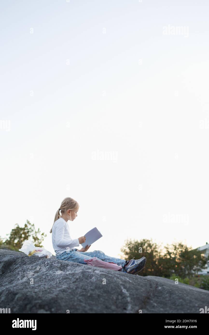 Vue latérale complète de la fille faisant ses devoirs pendant qu'elle est assise sur le rocher contre le ciel dégagé Banque D'Images