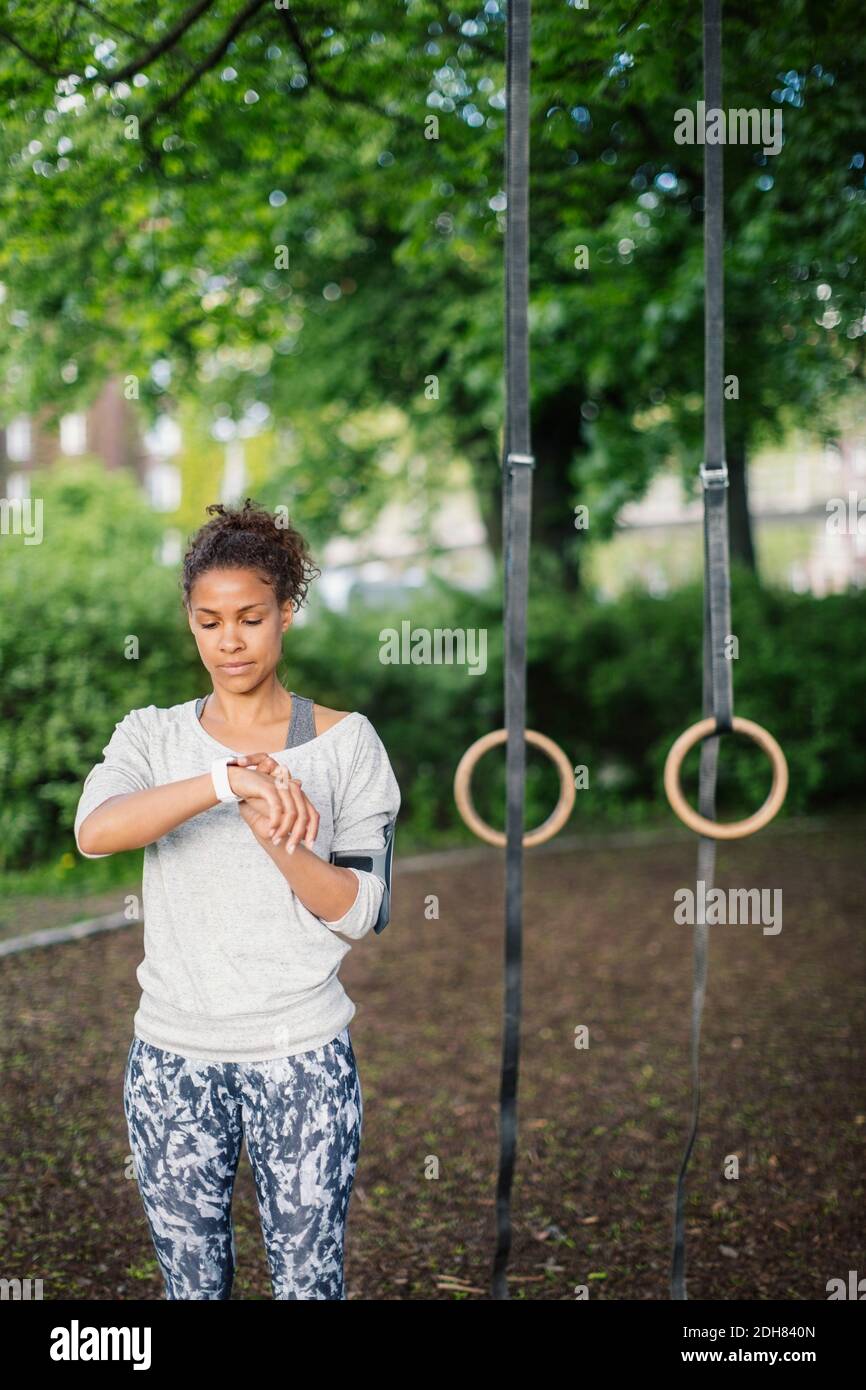 Femme utilisant une montre intelligente debout contre des anneaux de gymnastique dans le parc Banque D'Images