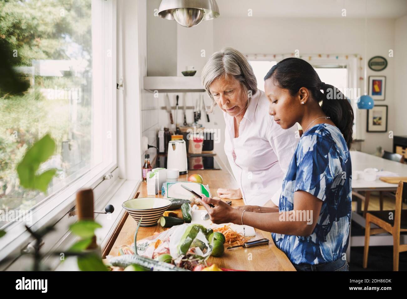 Femme âgée utilisant une tablette numérique avec une belle-fille tout en cuisinant de la nourriture dans la cuisine Banque D'Images
