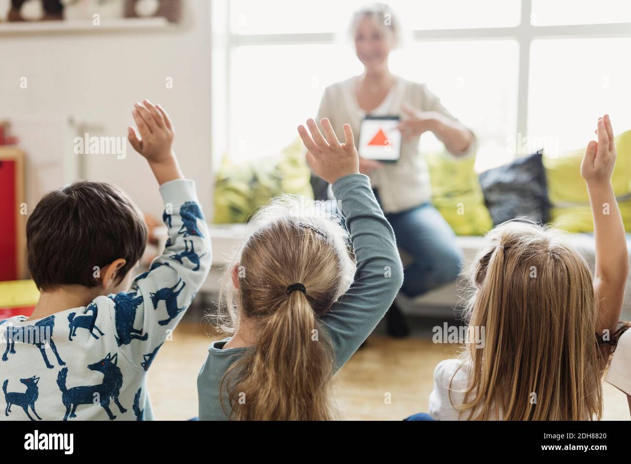 Vue arrière des élèves levant les mains pendant que le professeur affiche le numérique tablette à l'école maternelle Banque D'Images