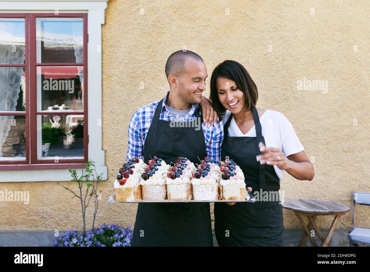 Des boulangers souriants tenant un plateau de pâtisseries tout en se tenant contre le mur café extérieur Banque D'Images