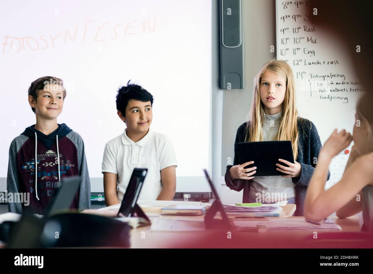 Bonne fille qui donne la présentation avec une tablette numérique parmi les étudiants dans salle de classe Banque D'Images