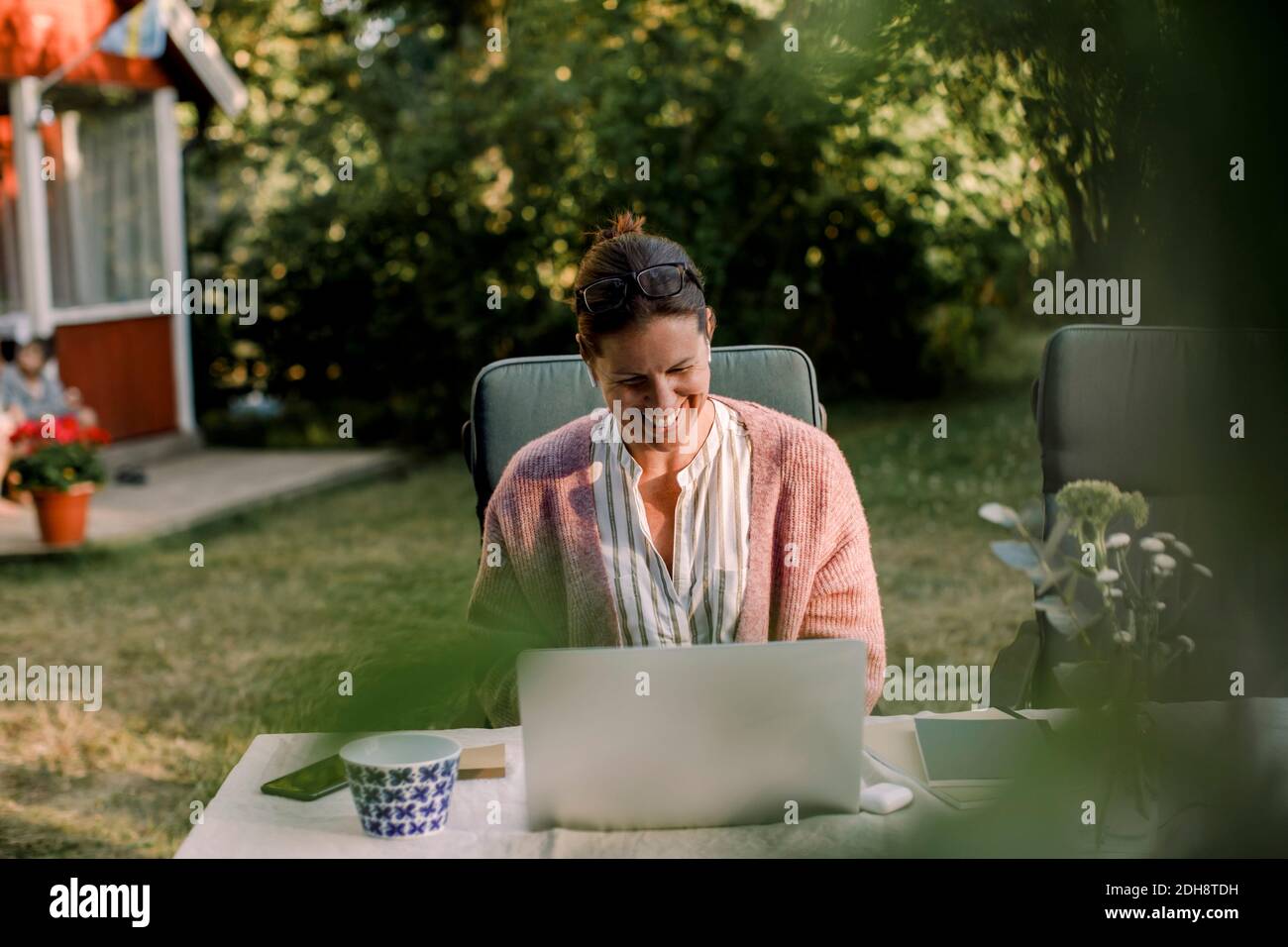 Femme souriante utilisant un ordinateur portable tout en étant assise à la table à manger jardin en été Banque D'Images