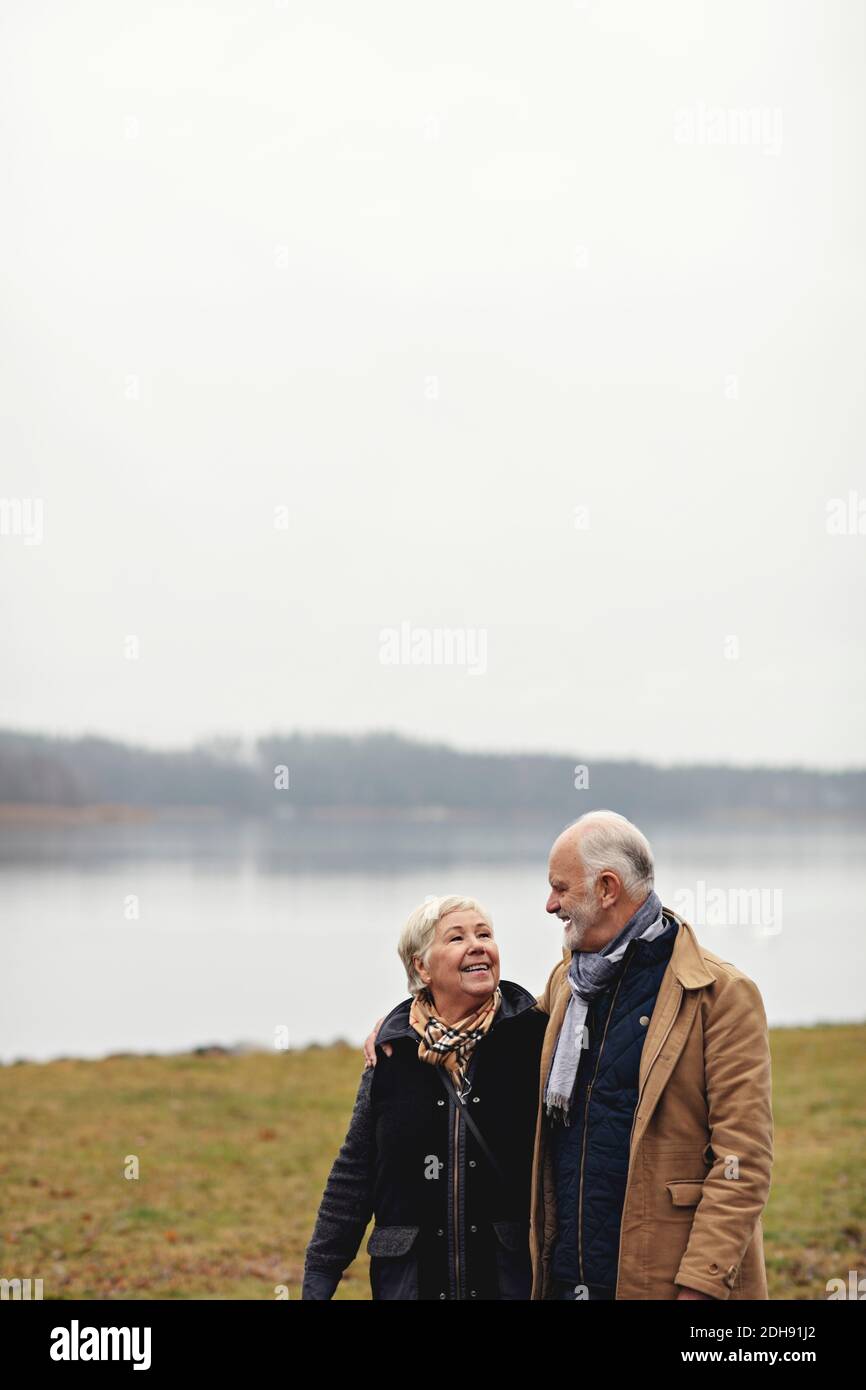 Couple senior souriant se regardant les uns les autres tout en restant debout lac contre ciel clair Banque D'Images