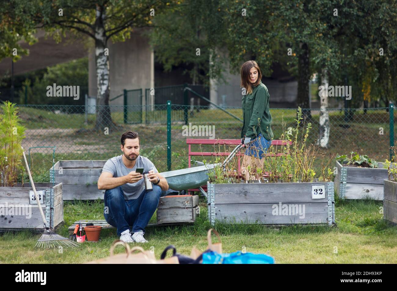 Homme de taille moyenne utilisant un téléphone portable pendant qu'une femme travaille jardin urbain Banque D'Images