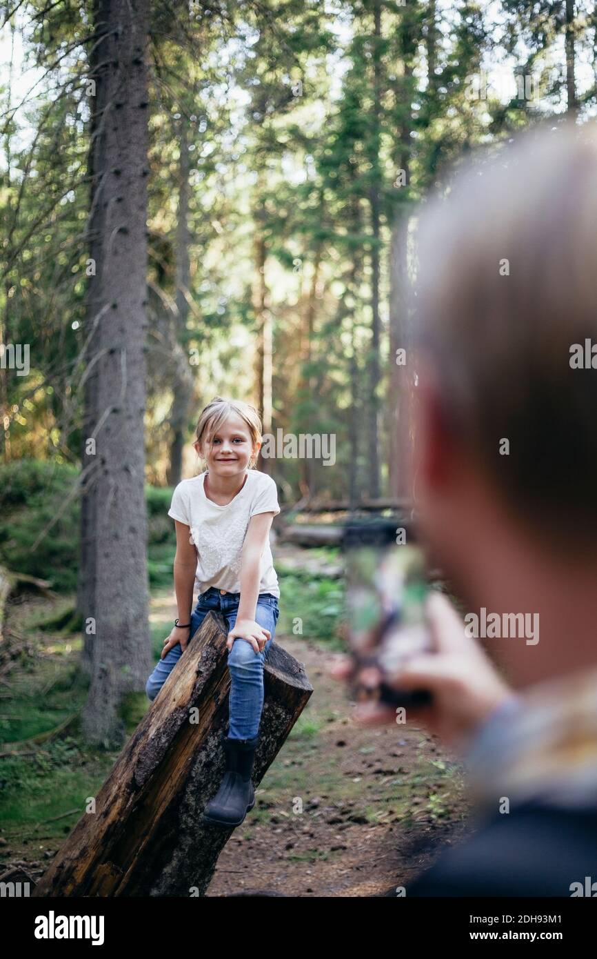 Fille souriante assise en rondins pendant que le père photographie avec intelligence téléphone en forêt Banque D'Images