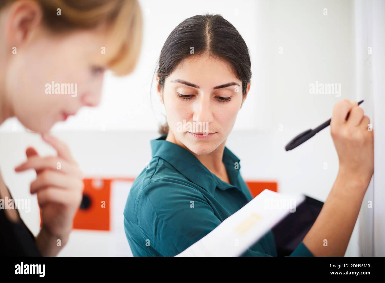 Femme d'affaires de taille moyenne, écrivant sur un tableau blanc pendant que son collègue lit le document au bureau Banque D'Images