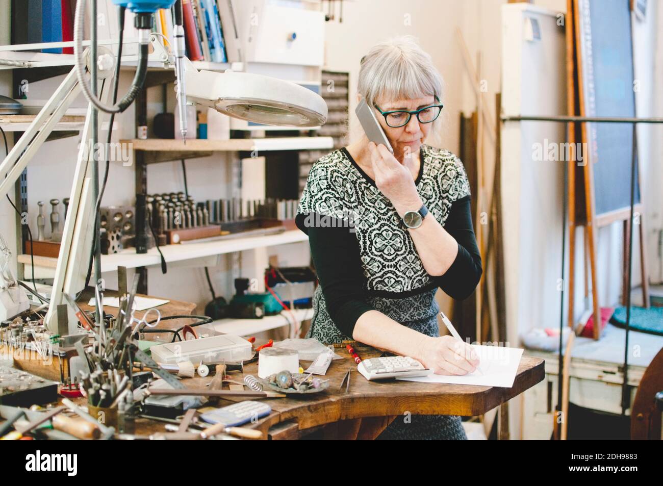 Femme âgée utilisant un téléphone portable tout en écrivant sur un document dans des bijoux atelier Banque D'Images