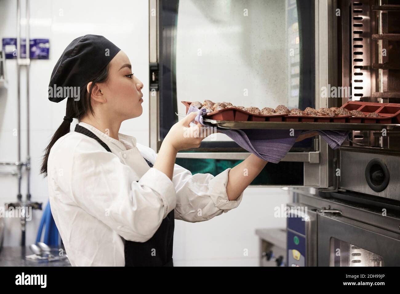 Vue latérale d'une femme chef en train de retirer un plateau de biscuits du four dans la cuisine commerciale Banque D'Images