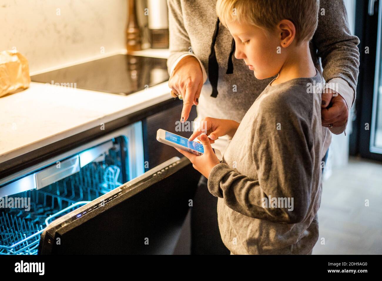 Un garçon apprend de sa mère tout en utilisant l'application mobile au lave-vaisselle dans la cuisine à la maison intelligente Banque D'Images
