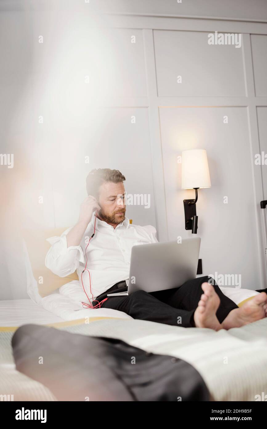 Homme d'affaires mature utilisant des écouteurs réglables pour ordinateur portable tout en étant assis sur le lit dans la chambre d'hôtel Banque D'Images