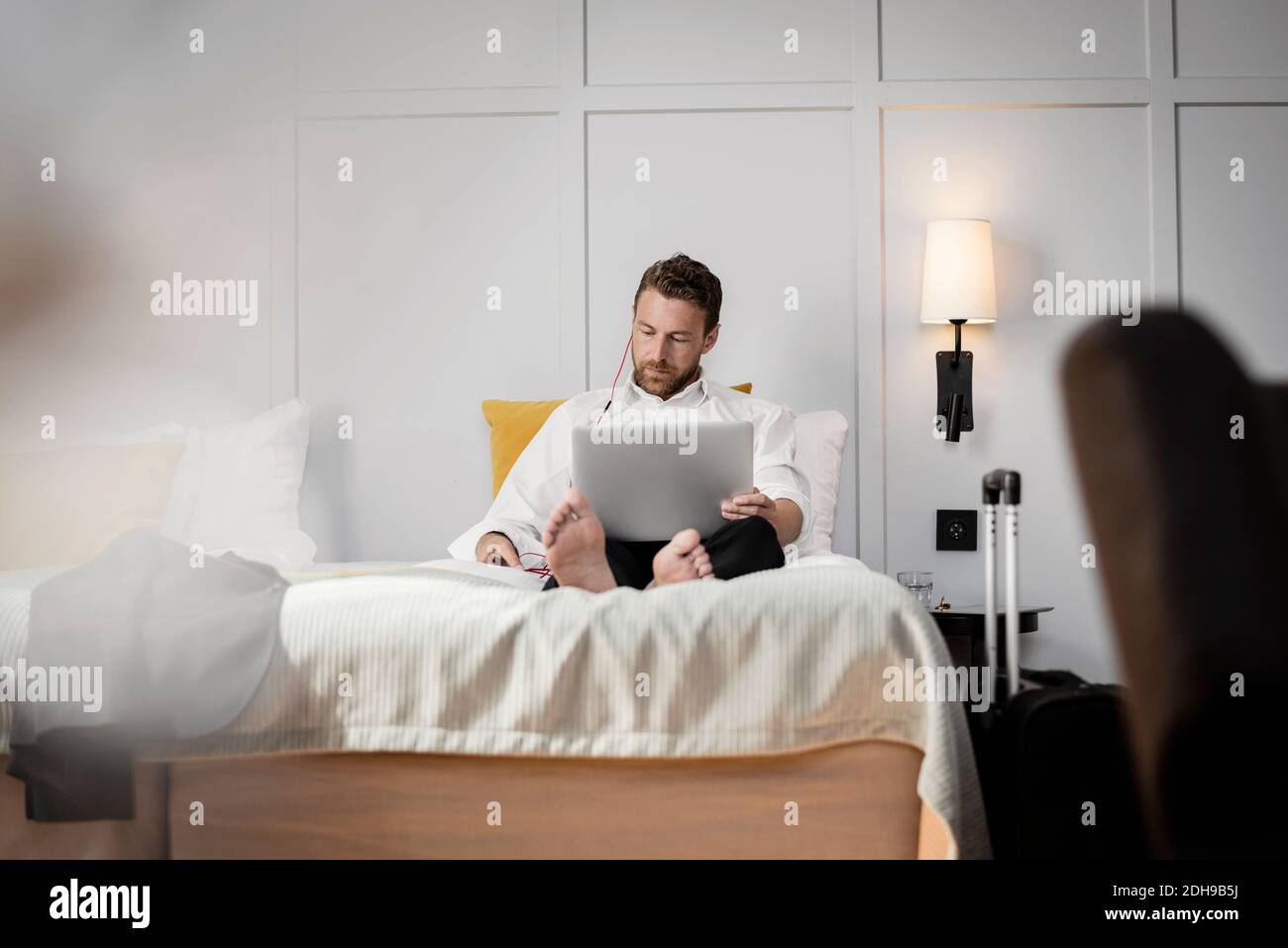 Homme d'affaires assis sur le lit en utilisant un ordinateur portable contre le mur dans l'hôtel chambre Banque D'Images