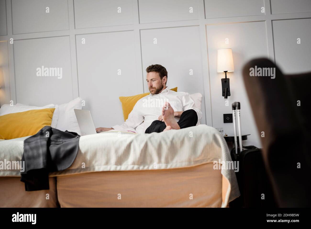 Homme d'affaires assis sur le lit en utilisant un ordinateur portable contre le mur dans l'hôtel chambre Banque D'Images