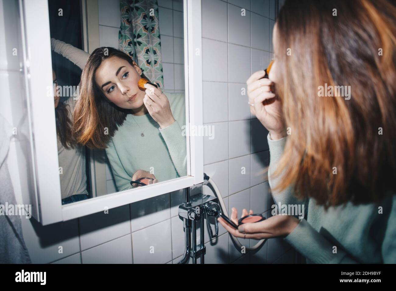 Jeune femme appliquant blush regardant dans le miroir à l'université dorm salle de bains Banque D'Images