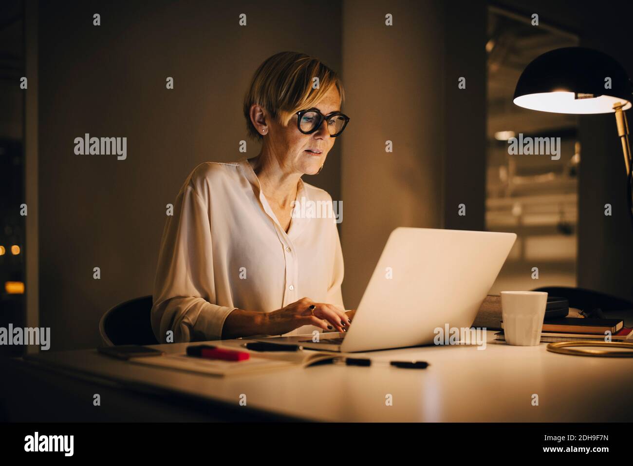 Femme d'affaires déterminée utilisant un ordinateur portable tout en travaillant tard dans la création éclairée lieu de travail Banque D'Images