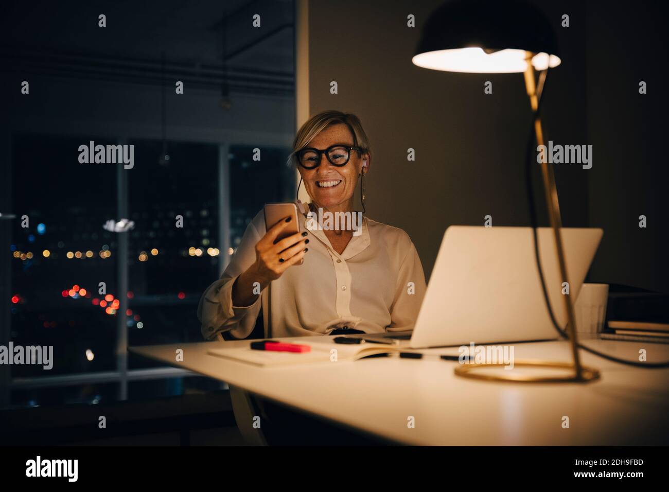 Femme d'affaires souriante utilisant un smartphone tout en étant assise avec un ordinateur portable à bureau éclairé dans un espace collègue Banque D'Images