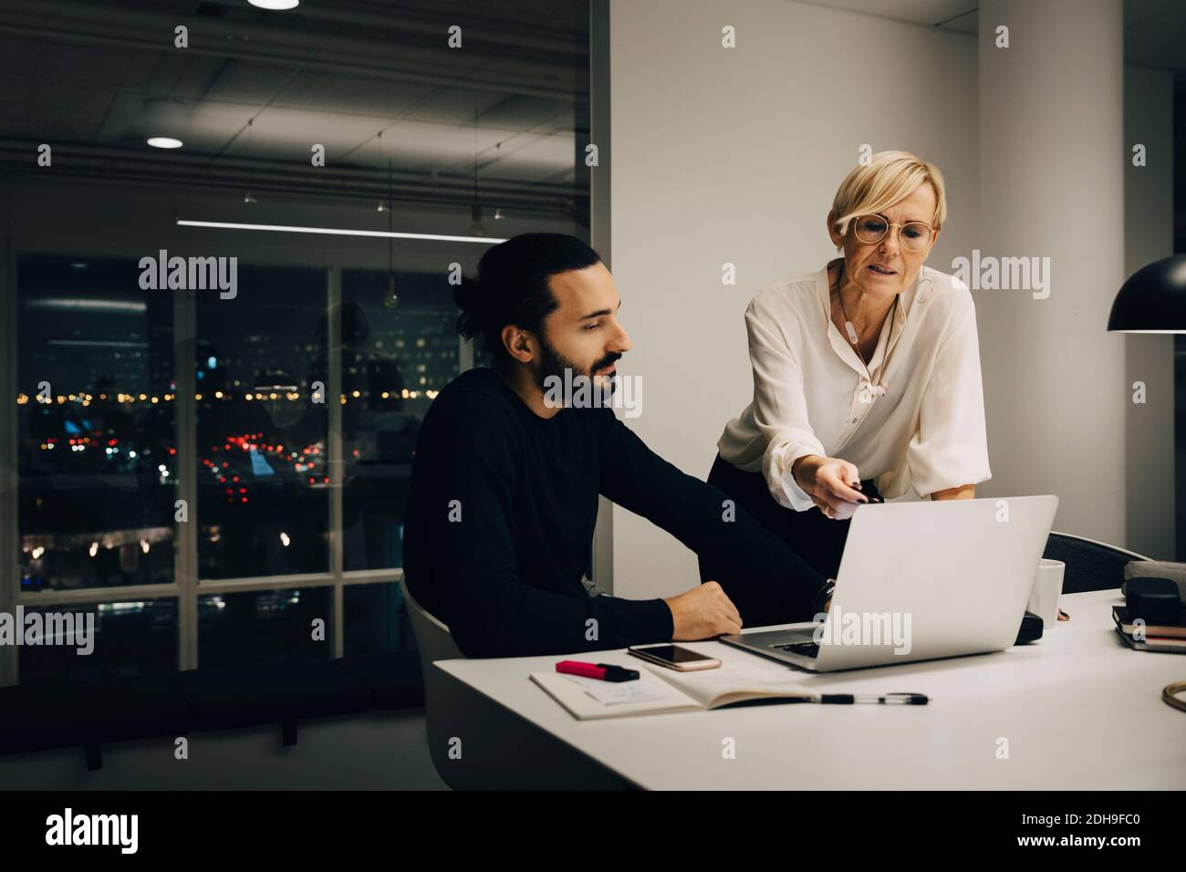 Les professionnels masculins et féminins planifient leur stratégie tout en discutant sur ordinateur portable dans un bureau éclairé Banque D'Images